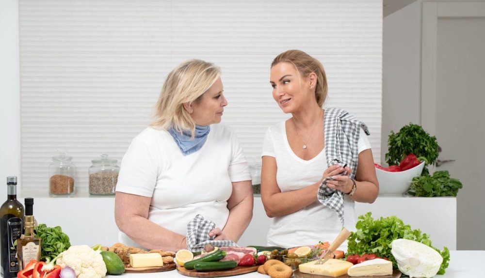 Two women in a kitchen preparing fresh vegetables and cheese with olive oil and spices.