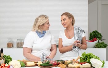 Two women in a kitchen preparing fresh vegetables and cheese with olive oil and spices.