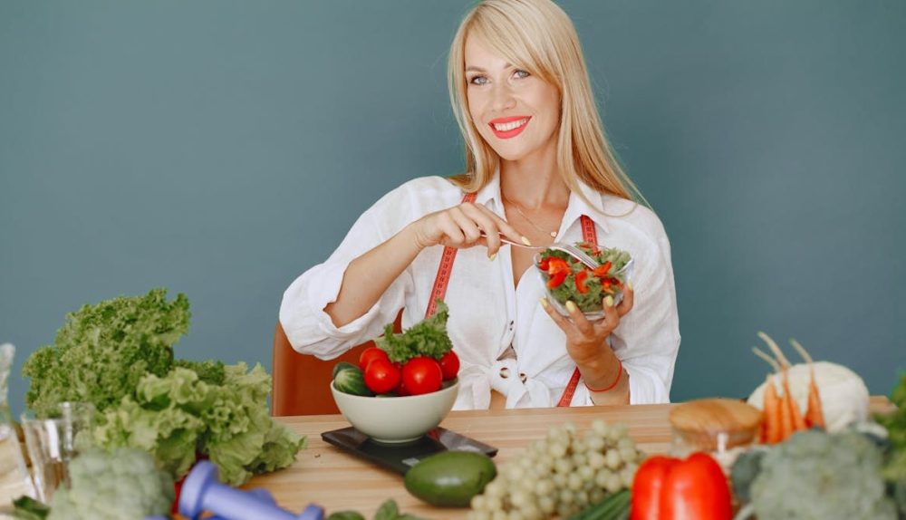 Blonde woman enjoying a fresh salad surrounded by vegetables, promoting healthy eating.